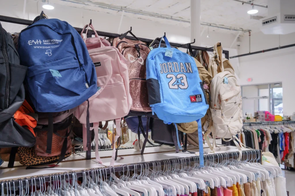 Assorted backpacks in blue, pink, gold, and neutral colors displayed above clothing racks inside Value World Barker Cypress.